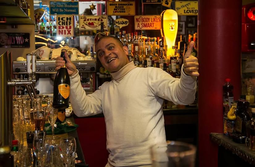 Cheffe Sherif hinter dem Tresen, lachend, mit einer Flasche in der Hand und erhobenem Daumen